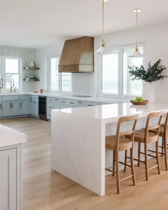 Ponte Vedra coastal kitchen with light blue cabinetry, brass hardware, a quartz waterfall island and a wood range hood