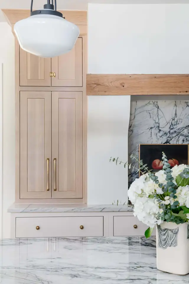Built-in cabinetry in natural oak with brass hardware beside a marble-clad fireplace and a schoolhouse pendant